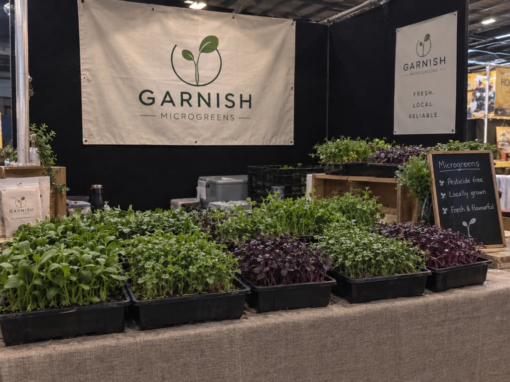 Garnish Microgreens market stall with trays of green and purple microgreens, branded banner, and a chalkboard highlighting pesticide-free locally grown produce.